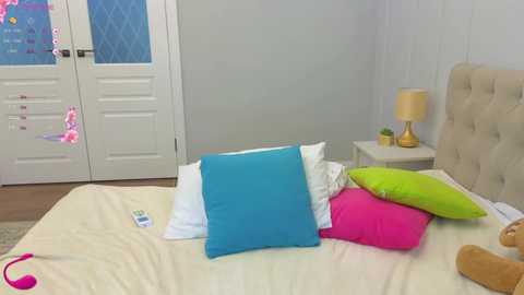 Video of a bright, modern bedroom with white walls, a beige tufted headboard, and colorful pillows on a cream bedspread. A closed white door with diamond-shaped glass panels is visible in the background.