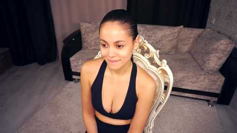Video of a young woman with medium brown skin and dark hair pulled back, wearing a black sports bra and black shorts, seated on a vintage white chair in a modern, dimly-lit living room.