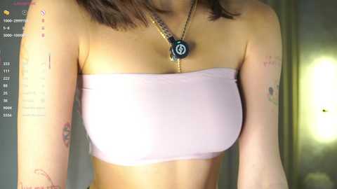Video of a woman with fair skin, medium brown hair, wearing a light pink strapless bandeau top, showcasing tattoos on her arms and a black pendant necklace. Background shows a dimly lit room with a window.