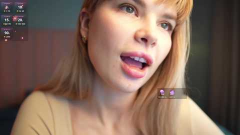 A close-up video of a Caucasian woman with long, blonde hair and light skin, wearing a beige top, smiling with her mouth open, taken indoors.