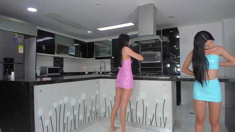 Video of two women with long black hair in tight, shiny pink and blue mini dresses, standing in a modern, minimalist kitchen with black countertops and stainless steel appliances.