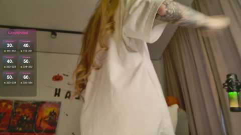 Video of a woman in a white nightgown, long red hair, tattoos on her arms, standing indoors with a digital display showing stats and a pumpkin.