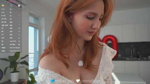 Video of a fair-skinned, red-haired woman in a white lace dress, standing in a modern kitchen with white cabinets and a red balloon.