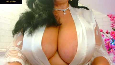 A video of a woman with long black hair, wearing a white satin robe that reveals her large, ample breasts, adorned with a delicate necklace. Background includes a white brick wall and a pink floral arrangement.