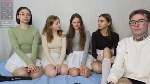 Media: Video of five young women and one man, all with light skin and straight hair, sitting on a bed in casual attire, against a plain white wall.