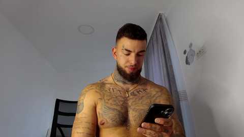 Video of a muscular, bearded man with tattoos covering his chest and arms, holding a smartphone, standing in a white-walled room with a grey curtain.