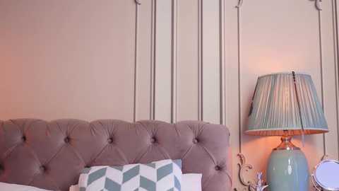Video of a modern bedroom with a beige tufted headboard, a green lamp with a striped lampshade, and a white bed with a blue and white geometric-patterned pillow.