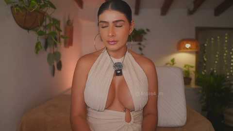 Video of a young, light-skinned Latina woman with dark hair in a bun, wearing a revealing, off-shoulder beige halter dress, adorned with large hoop earrings and a statement necklace, in a warmly lit, cozy room with green plants.