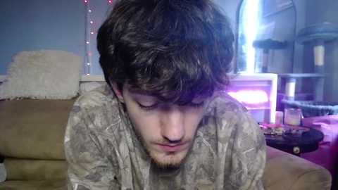 Video of a young man with short brown hair, wearing a light gray sweater, leaning forward, indoors with a beige couch, a cat bed, and bookshelves in the background.