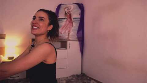 Video of a smiling, fair-skinned woman with brown hair in a ponytail, wearing a black tank top, standing in a dimly lit room with a wall mural of a woman in a pink dress.