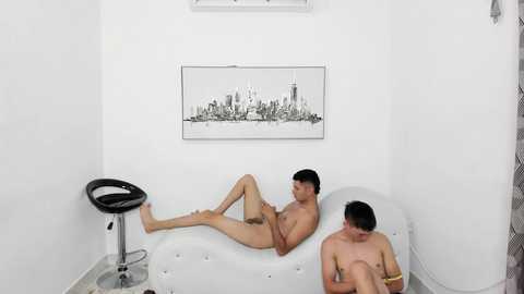 Video of two nude men, one sitting on a white chaise lounge, the other standing behind, in a minimalist white room with a cityscape print on the wall and a black stool.