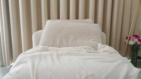 Media: Video of a neatly made bed with white sheets and pillows against beige curtains. A vase of pink and white flowers is on the right side.
