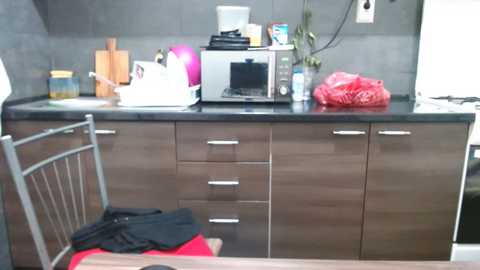 Video of a modern kitchen with dark wood cabinets, black countertops, and a microwave. A red plastic bag and a pink helmet are visible on the counter. A wooden chair with a black cloth is placed in the foreground.