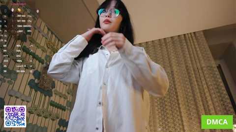 A video of a young Asian woman with long black hair, wearing glasses and a white lab coat, adjusting her collar. The background features a modern, beige-walled room with a floral-patterned curtain.