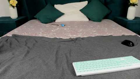 Video of a neatly made bed with a gray blanket, green pillows, a pink quilt, and a white keyboard on the floor.