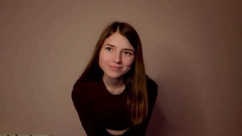 Video of a young Caucasian woman with long, straight brown hair, fair skin, and blue eyes, wearing a black long-sleeve top, smiling softly against a plain, light brown background.