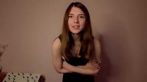 Video of a young woman with long brown hair, fair skin, and a slender physique, wearing a black dress, standing against a beige wall, arms crossed, smiling softly.