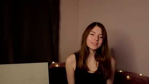 Video of a young woman with long brown hair, smiling, wearing a black top, sitting on a brown sofa against a beige wall with dark curtains and fairy lights.