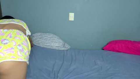 Video of a young woman in a bed, wearing yellow pajamas with pink patterns. She has a medium skin tone, and the room has a blue wall with a light switch.