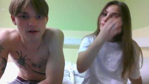 Media: Video of a shirtless, tattooed, muscular man with a shocked expression and a skinny, long-haired woman in a white shirt, both in a brightly lit bedroom.