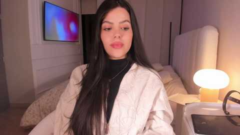 Video of a young woman with long black hair, fair skin, and full lips, wearing a white jacket over a black top, sitting on a bed with a white headboard. The room has a cozy, warm ambiance with a lit lamp and colorful wall art.