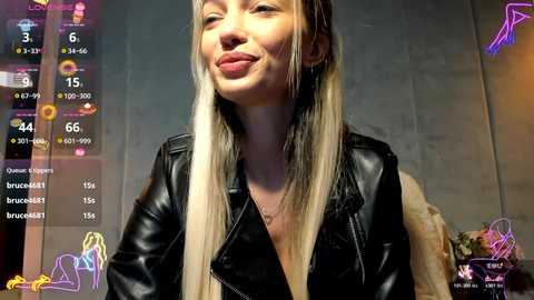 Video of a smiling Caucasian woman with long, straight blonde hair, wearing a black leather jacket, standing indoors with a textured concrete wall background.