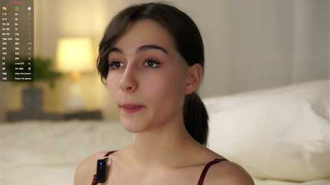 Media: A video of a young woman with olive skin and dark hair, wearing a red bralette, standing in a softly lit bedroom with a blurred background.