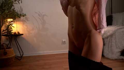 Video of a slender, topless woman with fair skin, wearing a pink-striped shirt, lowering black pants, in a dimly lit room with a wooden floor, potted plants, and a white fur rug.