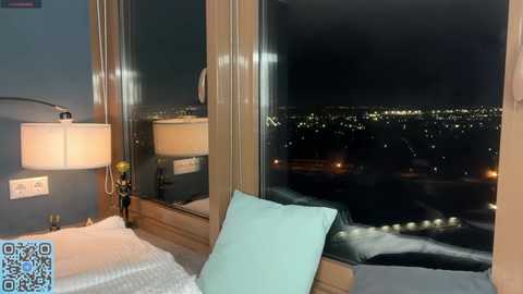 Video of a modern bedroom with a large window revealing a cityscape at night, featuring a turquoise pillow on a bed with white linens, a beige lamp, and a power outlet visible.
