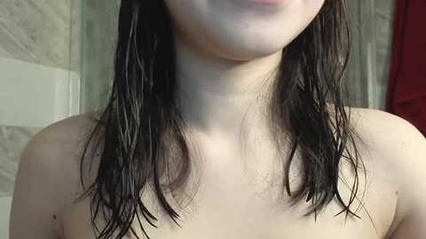 Video of a young, light-skinned woman with wet, dark brown hair, partially covering her breasts, standing in a bathroom with tiled walls and a red towel in the background.