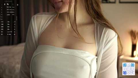 Video of a young woman with light skin and straight brown hair, wearing a white, see-through cardigan over a strapless top, smiling slightly. Background shows a bed with white sheets and a nightstand with a lamp. QR code in bottom right corner.