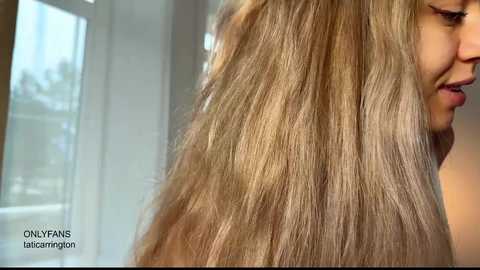 Video of a blonde woman with long, voluminous hair, smiling. The background shows a blurred indoor setting with large windows and natural light. Text on the image reads \"ONLYLYFANS TALCOSMELON.\