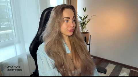 Video of a young woman with long, wavy blonde hair, wearing a light blue shirt, sitting in a black office chair in a bright room with a green plant and window.