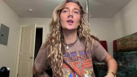 Video of a young woman with long, wavy blonde hair, wearing a brown Nirvana t-shirt, silver hoop earrings, and a nose ring, in a dimly lit room with beige walls and a door.