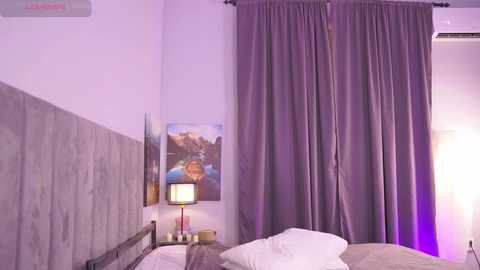 A video of a modern bedroom with a lavender headboard, white bedding, a wooden nightstand, a framed landscape painting, and a floor lamp with a purple shade.