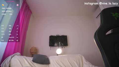 Video of a dimly lit, minimalist bedroom with white walls, a pink curtain, a black wall-mounted plant, and a gaming chair.