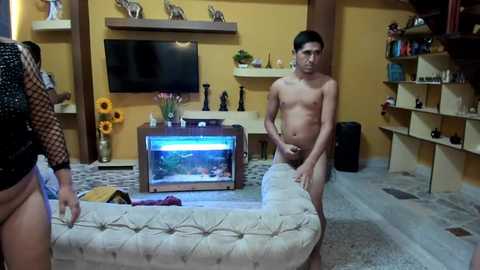Video of a modern living room with a naked young man standing by a fish tank, a woman in a black top and shorts, and decorative shelves.