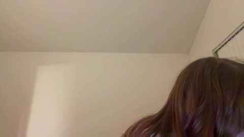 A video shows the side profile of a woman with long, wavy brown hair, standing against a plain, light beige wall. The image captures a candid, slightly blurred moment.