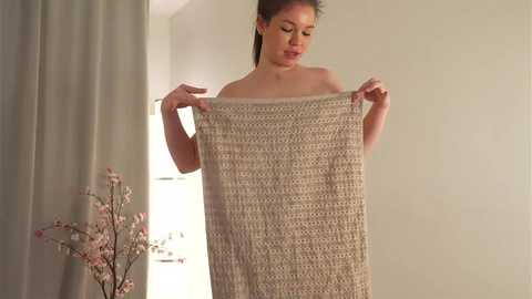 Media: Video of a woman with light brown skin and long, dark hair, standing in a softly lit room, holding a beige, knitted blanket. Behind her, a vase of pink cherry blossoms and white curtains are visible.
