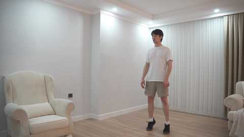 Video of a young Asian man with short dark hair, wearing a white t-shirt, beige shorts, white socks, and black shoes, standing in a minimalist, modern living room with white walls, beige armchairs, and vertical blinds.
