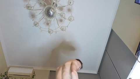 A video of a man with short brown hair, partially blurred, standing in a modern living room with beige walls and a white ceiling fan.