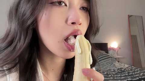 Video of an Asian woman with long black hair, light skin, and full lips, eating a peeled banana, in a bedroom with a bed, nightstand, and lamp.