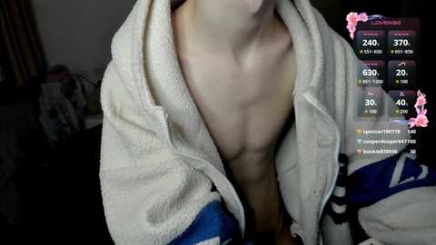 Video of a shirtless, fair-skinned man with a muscular build, wearing a white towel draped around his shoulders. Background includes a cluttered room with a bed, and a smartphone screen displaying fitness app data.