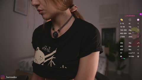 Media: Video of a young woman with light skin and auburn hair, wearing a black t-shirt with a cartoon whale design. Background features a dimly lit room with blurred furniture. Social media app interface overlaid.