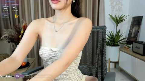 A video of an East Asian woman with fair skin and dark hair in a strapless, sequined dress, sitting at a desk in a modern room.