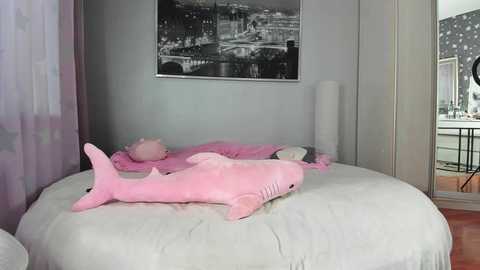 A video of a minimalist bedroom with a white bed featuring two pink dolphin plush toys and a framed black-and-white cityscape above. The room has soft lighting, with sheer curtains on the left and a tall white lamp on the right.