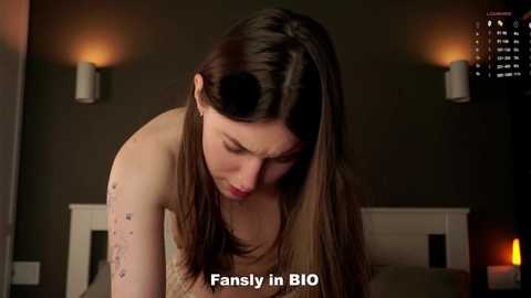 Video of a nude woman with long brown hair, leaning forward, in a dimly lit room with two wall-mounted lamps and a lit candle. Text reads \"Fansly in BIO.\