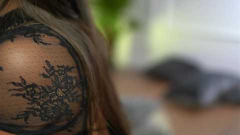 Media: A close-up video of a woman's bare shoulder adorned with intricate black lace, showing detailed floral patterns. The background is blurred, featuring a green plant and white walls.
