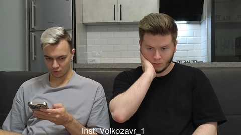 Video of two young men with short hair, one in gray, the other in black, sitting on a gray sofa in a modern kitchen, the man in black holding a phone, looking thoughtful.