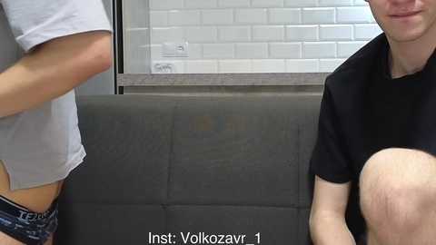 A video of two young people in a modern setting: one person wearing a white shirt and blue shorts, and the other in a black shirt. Background includes white subway tiles and a gray counter.